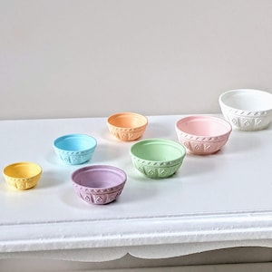 Peut inclure: Un ensemble de sept bols à mélanger de couleur pastel, allant du petit au grand. Les bols sont en céramique et ont un design texturé et gaufré. Les bols sont disposés en rangée sur une surface blanche.