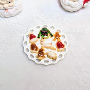 Peut inclure: Une assiette blanche décorative contient des biscuits miniatures sur le thème de Noël, dont un bonnet de Père Noël, une mitaine et un renne. Les biscuits sont décorés de glaçage rouge, vert et marron. Deux figurines de Père Noël sont en arrière-plan.