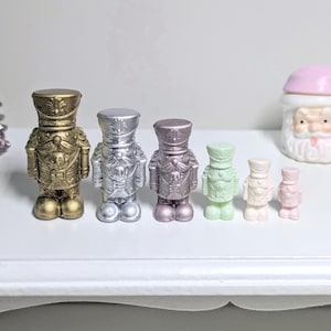 Puede incluir: Una colección de figuras de cascanueces en miniatura en varios colores metálicos, incluyendo dorado, plateado y malva, dispuestas sobre una superficie blanca. Decoraciones navideñas adicionales incluyen una figura de Papá Noel y un árbol de Navidad brillante.