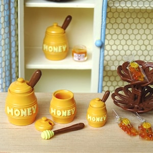 Peut inclure: Un ensemble de pots de miel miniatures avec des louches en bois, une louche à miel et un support à deux étages avec des sachets de bonbons au miel. Les pots de miel sont jaunes avec le mot "HONEY" imprimé dessus.