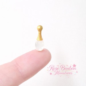 Pode incluir: Miniatura de frasco de perfume com tampa dourada e corpo transparente em forma de pêra. O frasco é segurado na ponta de um dedo. O texto "Rose Bonbon Miniatures" está no canto inferior direito.