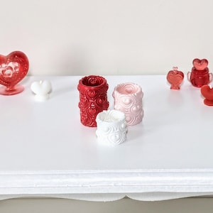 May include: A selection of Valentine's Day decorations. The image shows red, pink, and white heart-shaped candles, candle holders, and decorative glass pieces. The candle holders have heart patterns. The items are arranged on a white surface.