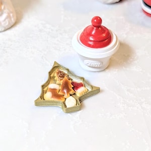 Peut inclure: Un plateau doré en forme de sapin de Noël rempli de bonbons festifs. Les bonbons comprennent une chaussette, un arbre et une gâterie rouge et blanche. Un récipient blanc avec un couvercle rouge et le mot "CANDY" est également présent.