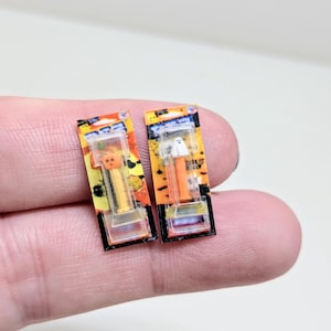 Puede incluir: Dos dispensadores PEZ en miniatura con temática de Halloween. Uno presenta una calabaza, el otro un fantasma. Ambos son naranjas y blancos, con embalaje colorido. Los dispensadores se sostienen entre los dedos.