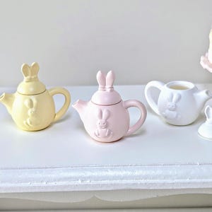 May include: A selection of miniature ceramic teapots and a bunny-shaped lid. The teapots are in pastel yellow, pink, and white. Each teapot features a bunny design. A small cake and a larger cake are in the background.