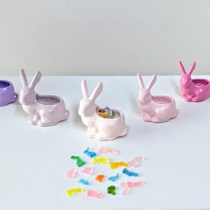 May include: Five ceramic bunny planters, four are pink and one is purple. The planters are filled with colourful candy pieces.
