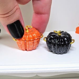 Può includere: Due pentole in miniatura a forma di zucca, una arancione con macchie bianche e una nera con macchie bianche. Entrambe hanno manici dorati.
