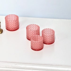 Peut inclure: Quatre bougeoirs en verre rose avec un motif en forme de cœur texturé. Les bougeoirs sont disposés en rangée sur une surface blanche.