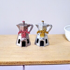 Peut inclure: Deux cafetières miniatures en argent avec un design en forme de maison. Les cafetières portent l'inscription "Mocha Latte" en lettres dorées et rouges.