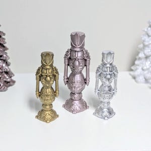 Könnte beinhalten: Eine Sammlung dekorativer Weihnachtsfiguren, darunter drei Nussknacker in Gold, Silber und Mauve. Zwei glitzernde Weihnachtsbäume und andere weihnachtliche Ornamente sind ebenfalls vorhanden.