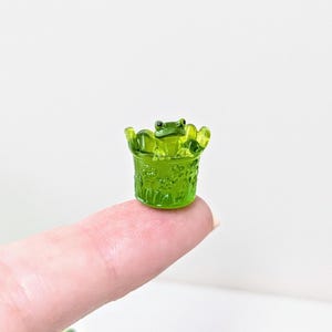 Könnte beinhalten: Eine kleine, durchscheinende grüne Froschfigur in einem passenden grünen Topf. Der Frosch hat große Augen und sitzt im Topf. Der Topf hat ein Blumenmuster. Die Figur wird auf einem Finger vor weißem Hintergrund gehalten.