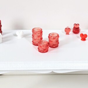 Peut inclure: Trois bougeoirs en verre rouge avec un motif en forme de cœur. Les bougeoirs sont de tailles différentes et sont disposés sur une surface blanche.