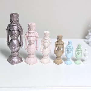 Peut inclure: Une collection de figurines casse-noisettes décoratives de différentes tailles et couleurs, dont argent, rose, blanc et marron. Les figurines sont disposées en rangée, mettant en valeur leurs motifs détaillés et leur aspect festif.