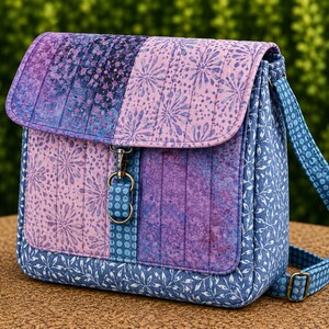 May include: A quilted fabric crossbody bag featuring a patchwork design in shades of purple, lavender, and blue. The bag has a flap closure with a metal swivel clasp, a matching adjustable shoulder strap, and various floral and geometric patterns.