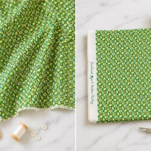 May include: A green fabric with a repeating oval pattern and a white selvage edge printed with the text Freshcut by Heather Bailey. The fabric is displayed alongside gold-toned scissors, small white buttons, and wooden thread spools.