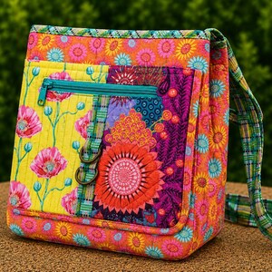 May include: A quilted crossbody bag featuring a vibrant floral pattern in shades of orange, yellow, pink, and teal. The bag includes a front zipped pocket with a teal zip, a plaid fabric strap, and two bronze-coloured metal rings on the front.