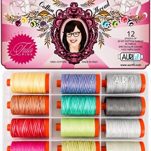 Op de afbeelding: Een rechthoekige doos met 12 klossen 50 WT katoengaren in diverse kleuren. Het deksel toont een portret van een persoon met een bril, de tekst Premium Cotton Thread, Tula Pink en Aurifil, met vermelding van 1422 yards, gemaakt in Italië.