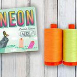 Puede incluir: Una caja rectangular etiquetada NEON Limited Edition AuriFil con una ilustración de carretes de hilo sobre una roca, junto a tres carretes grandes de hilo de coser en colores vibrantes naranja, verde lima y rosa brillante.