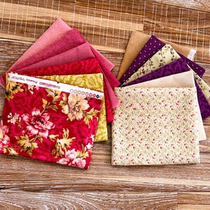 Op de afbeelding: Assortiment van stoffen vierkanten in verschillende kleuren en patronen. Inclusief bloemen-, stippen- en effen ontwerpen in tinten roze, rood, geel, paars en beige. De stof is gerangschikt op een houten oppervlak.