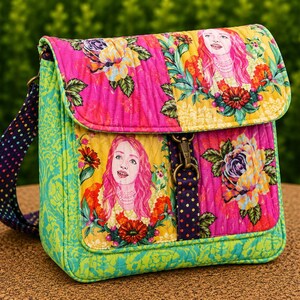 May include: A quilted crossbody bag featuring a vibrant floral pattern with pink and yellow sections. The design includes illustrations of a person with long pink hair surrounded by flowers. It has a dark strap with colourful polka dots and a metal clasp.