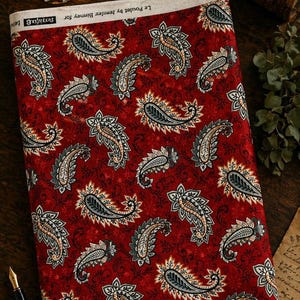 May include: A folded fabric featuring a red backdrop adorned with a repeating paisley design in grey, cream, and black. The fabric is labelled "Le Poulet by Jennifer Bimney for".
