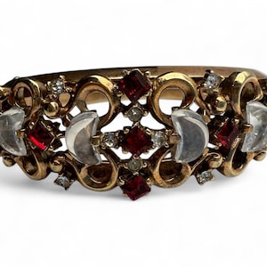 Puede incluir: Un brazalete de tono dorado con un diseño delicado e intrincado que presenta diamantes de imitación rojos y transparentes. El brazalete tiene una forma curva y está adornado con una variedad de elementos decorativos.