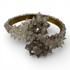 May include: A vintage bracelet with a gold-tone metal band and clusters of clear and smoky gray beads. The beads are faceted and arranged in floral-like patterns, creating a glamorous accessory.