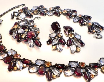 Può includere: Un set di gioielli vintage composto da una collana, un braccialetto e orecchini. Il set è realizzato in metallo color argento e presenta strass trasparenti, viola e ambra. La collana e il braccialetto sono entrambi di stile a catena con chiusura a clip. Gli orecchini sono di stile clip.