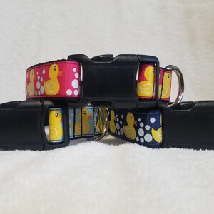 May include: Three dog collars with a rubber ducky pattern. The collars are in pink, blue, and navy blue. Each collar has a black plastic buckle and a silver D-ring.