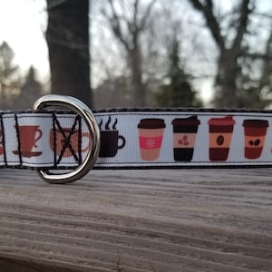 May include: A white dog collar with a black buckle and a silver D-ring. The collar features a repeating pattern of coffee cups and coffee to-go cups in various colors and designs.