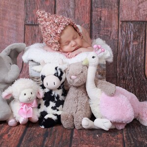 May include: A sleeping newborn wearing a pink and cream knitted hat, resting on a white fluffy surface. Soft toys of an elephant, lamb, cow, bunny, and a pink and white ostrich surround the baby, creating a cosy scene.