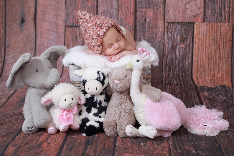 May include: A sleeping newborn wearing a pink and cream knitted hat, resting on a white fluffy surface. Soft toys of an elephant, lamb, cow, bunny, and a pink and white ostrich surround the baby, creating a cosy scene.