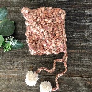 May include: A hand-knitted baby bonnet in shades of pink, cream, and mauve. The bonnet has a chunky knit texture and two cream-coloured pom-poms attached to the ends of the ties. The bonnet is designed to keep a baby's head warm.