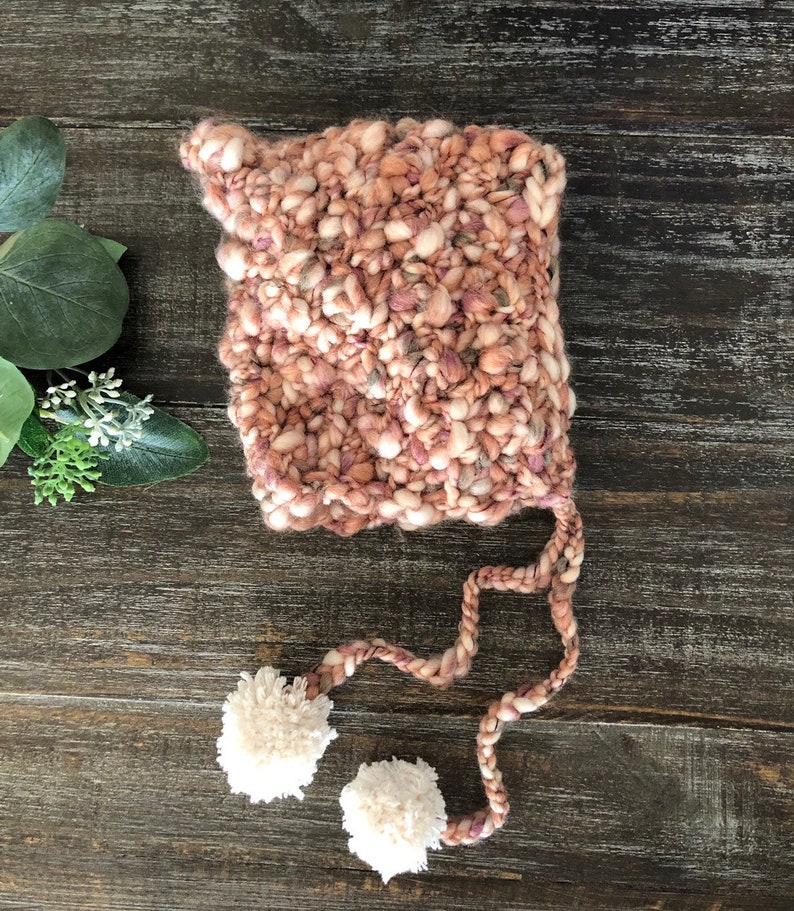 May include: A hand-knitted baby bonnet in shades of pink, cream, and mauve. The bonnet has a chunky knit texture and two cream-coloured pom-poms attached to the ends of the ties. The bonnet is designed to keep a baby's head warm.