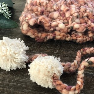 May include: A close-up of a hand-knitted hat in shades of pink, cream, and brown. The hat features a thick, textured yarn and is adorned with two fluffy cream-coloured pom-poms attached by a braided cord. A cosy winter accessory.