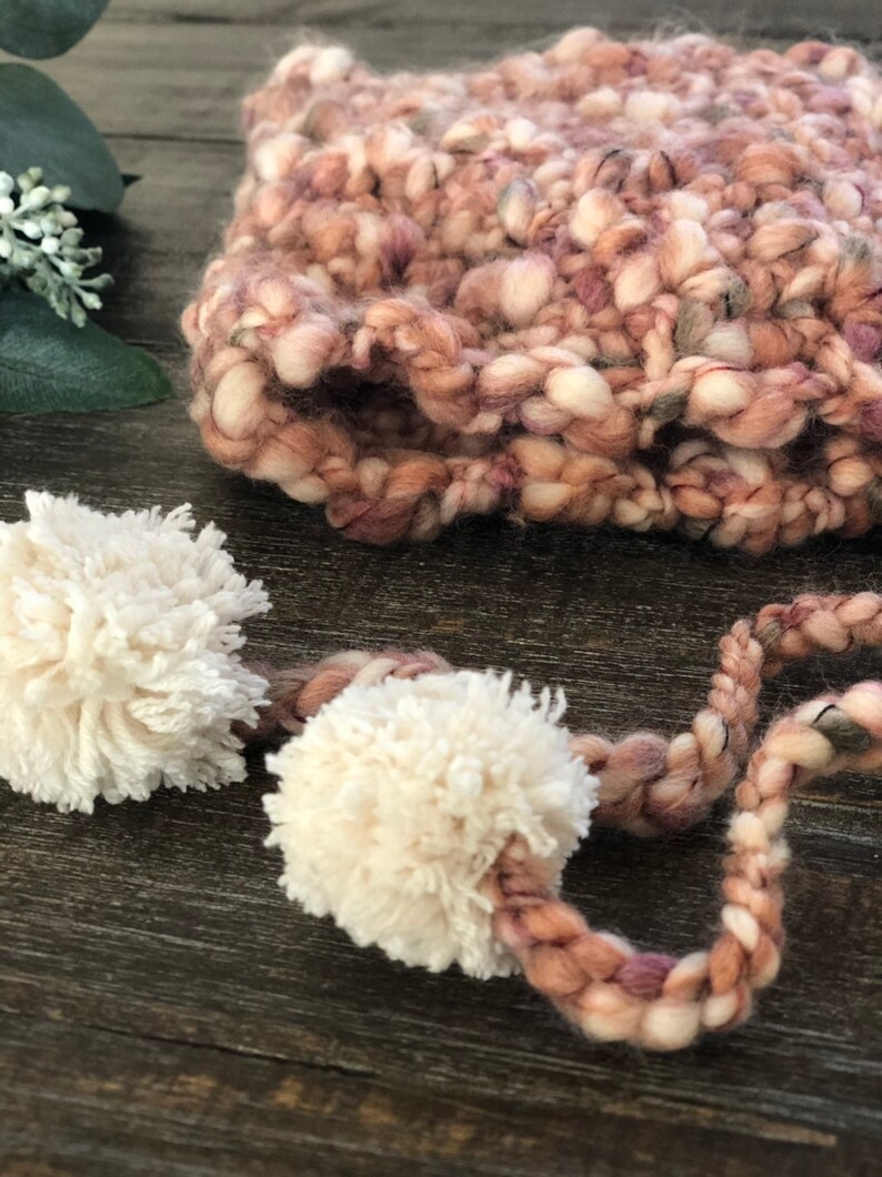 May include: A close-up of a hand-knitted hat in shades of pink, cream, and brown. The hat features a thick, textured yarn and is adorned with two fluffy cream-coloured pom-poms attached by a braided cord. A cosy winter accessory.