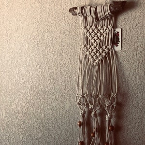 May include: A handmade macrame wall hanging. The off-white cotton fiber is knotted into a diamond pattern and hangs from a piece of driftwood. Wooden beads are attached to the bottom. A small tag is attached to the right side.