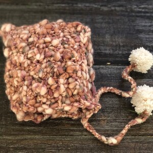 May include: A chunky-knit hat in pink, beige, and grey, with a textured appearance. Two white pom-poms are attached by a braided cord. The hat is designed for warmth and style, a cozy accessory for cooler weather.