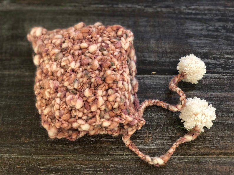 May include: A chunky-knit hat in pink, beige, and grey, with a textured appearance. Two white pom-poms are attached by a braided cord. The hat is designed for warmth and style, a cozy accessory for cooler weather.