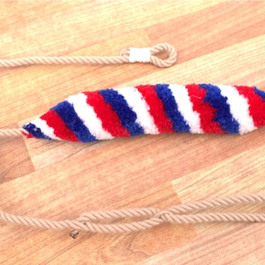 Może przedstawiać: Torba do procy w paski czerwono-biało-niebieskie z przymocowaną beżową sznurówką.