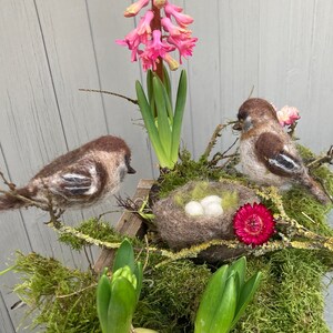 May include: Two brown and white felt birds perched on a branch with a nest of three white eggs. The nest is made of brown felt and is surrounded by green moss and a pink flower. The birds are perched on a branch with green leaves and a pink flower.