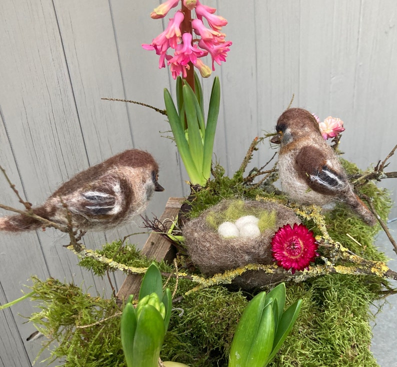 May include: Two brown and white felt birds perched on a branch with a nest of three white eggs. The nest is made of brown felt and is surrounded by green moss and a pink flower. The birds are perched on a branch with green leaves and a pink flower.