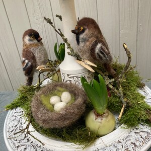 May include: A white ceramic vase with a brown felt nest containing three white eggs. Two brown and white felt birds are perched on a branch with green leaves. The vase and nest are surrounded by green moss and a green hyacinth bulb.