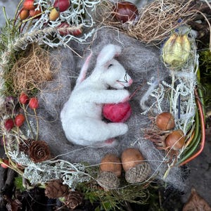 May include: A handmade felted white bunny cradling a red heart, nestled in a wreath of natural elements. The wreath includes dried berries, pinecones, acorns, and moss, creating a rustic, woodland theme.