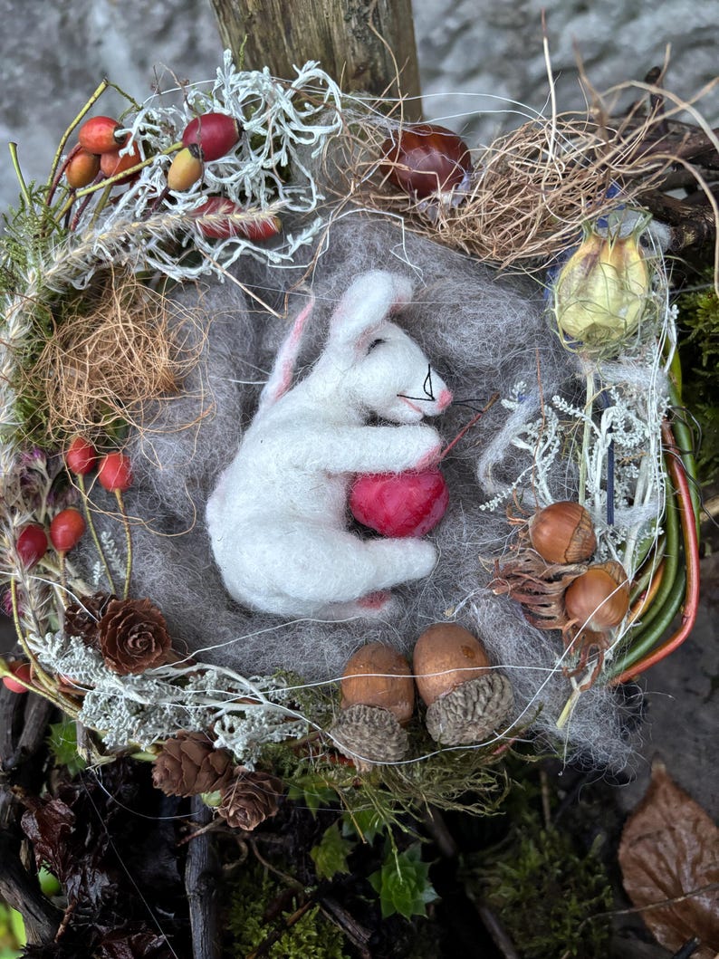 May include: A handmade felted white bunny cradling a red heart, nestled in a wreath of natural elements. The wreath includes dried berries, pinecones, acorns, and moss, creating a rustic, woodland theme.