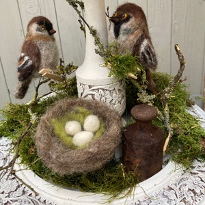 May include: A white ceramic pedestal with a brown and white bird perched on a branch. A small nest with three white eggs sits on the pedestal, surrounded by green moss. A rusty metal weight sits on the pedestal.