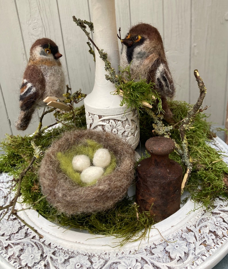 May include: A white ceramic pedestal with a brown and white bird perched on a branch. A small nest with three white eggs sits on the pedestal, surrounded by green moss. A rusty metal weight sits on the pedestal.