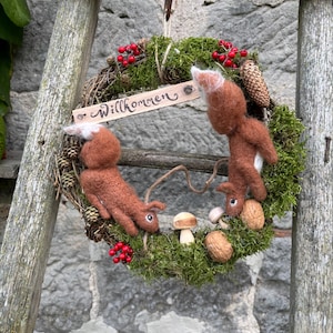 May include: A circular wreath made of twigs and green moss, decorated with two needle-felted brown squirrels, red berries, pinecones, walnuts, and a small mushroom. A wooden sign with the word Willkommen is attached to the top.