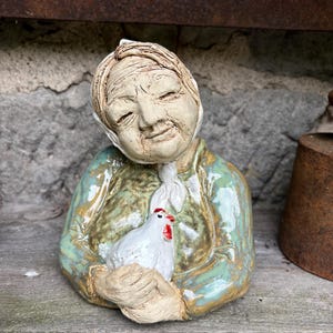 Könnte beinhalten: Eine Keramikfigur einer älteren Person mit weißer Kopfbedeckung, die ein grün und braun glasiertes Hemd trägt und ein kleines weißes Huhn mit rotem Kamm hält. Die Skulptur zeichnet sich durch detaillierte Gesichtsstrukturen und Glanz aus.