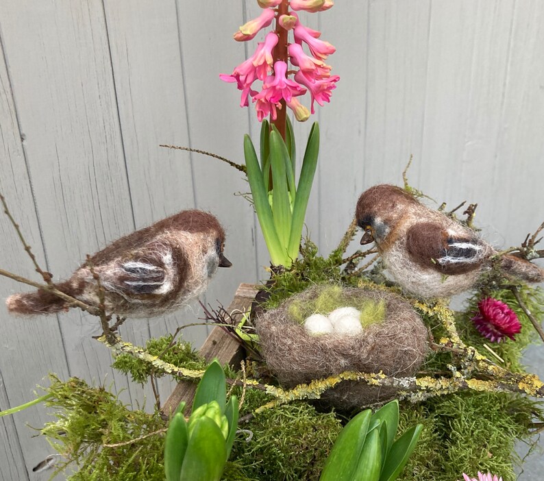 May include: Two brown and white felt birds perched on a nest with three white eggs. The nest is surrounded by green moss and a pink hyacinth flower.